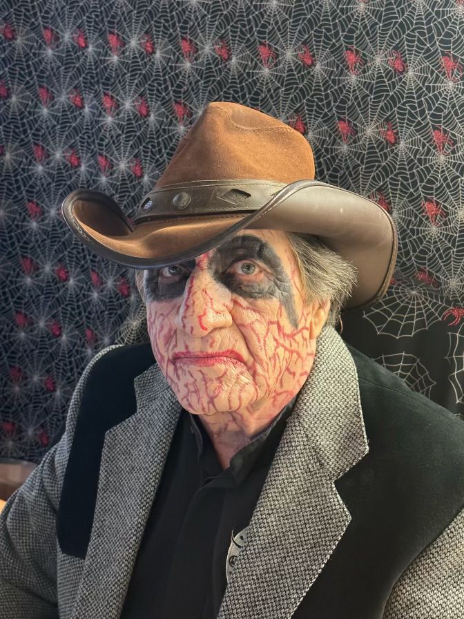 Dr. Ghoul in a cowboy hat and veiny face makeup in front of spiderweb wallpaper.